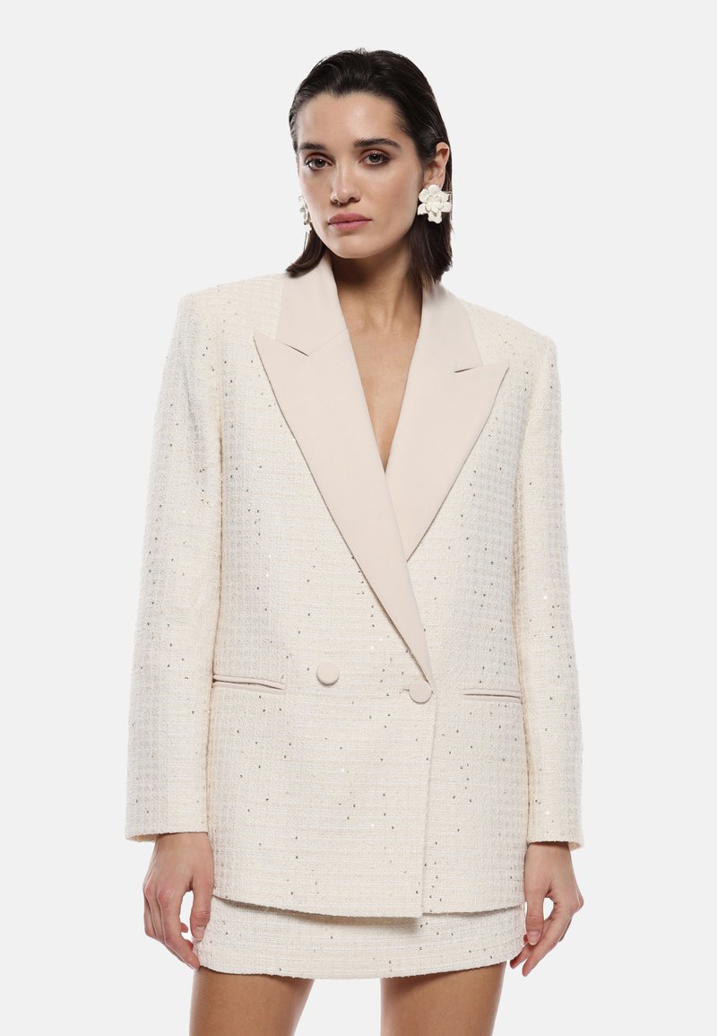 Blazer in tweed beige con un elegante risvolto chiaro, design doppiopetto e sottili accenti metallici, abbinato a una gonna corta coordinata.