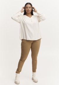 Blusa crema con scollo a V e dettagli con bottoni, abbinata a pantaloni beige aderenti e stivaletti bianchi alla caviglia. Texture liscia e vestibilità rilassata.