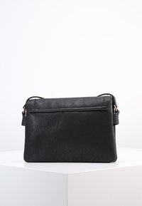 Borsa a tracolla in pelle nera con pattina, forma rettangolare e tracolla regolabile, posizionata su una piattaforma bianca.