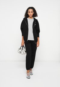 Felpa nera con zip, t-shirt grigia, pantaloni jogger neri e scarpe flat argentate. Una piccola borsa PUMA bianca e nera con manici corti.