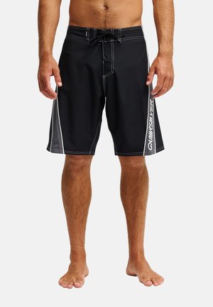 Mężczyzna w czarnych szortach do surfingu z szarymi bocznymi panelami i białym logo Quiksilver na prawej nodze, stojący boso na jednolitym tle.