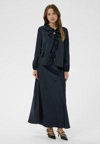 Marineblauwe satijnen blouse met ruches aan de voorkant en kraag, gecombineerd met een bijpassende lange rok. De outfit heeft een gladde textuur en een vloeiend ontwerp.