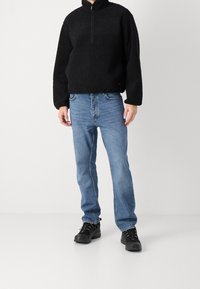 Persona che indossa un pile nero con mezza cerniera, jeans blu e scarpe da trekking nere, in piedi davanti a uno sfondo bianco uniforme.