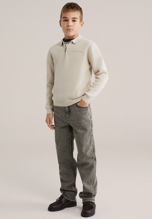 Svetlo béžový pulóver so zipsovým golierom, textúrovaná tkanina, sivé džínsy a tmavohnedé topánky. Pulóver má malé vrecko na hrudi.