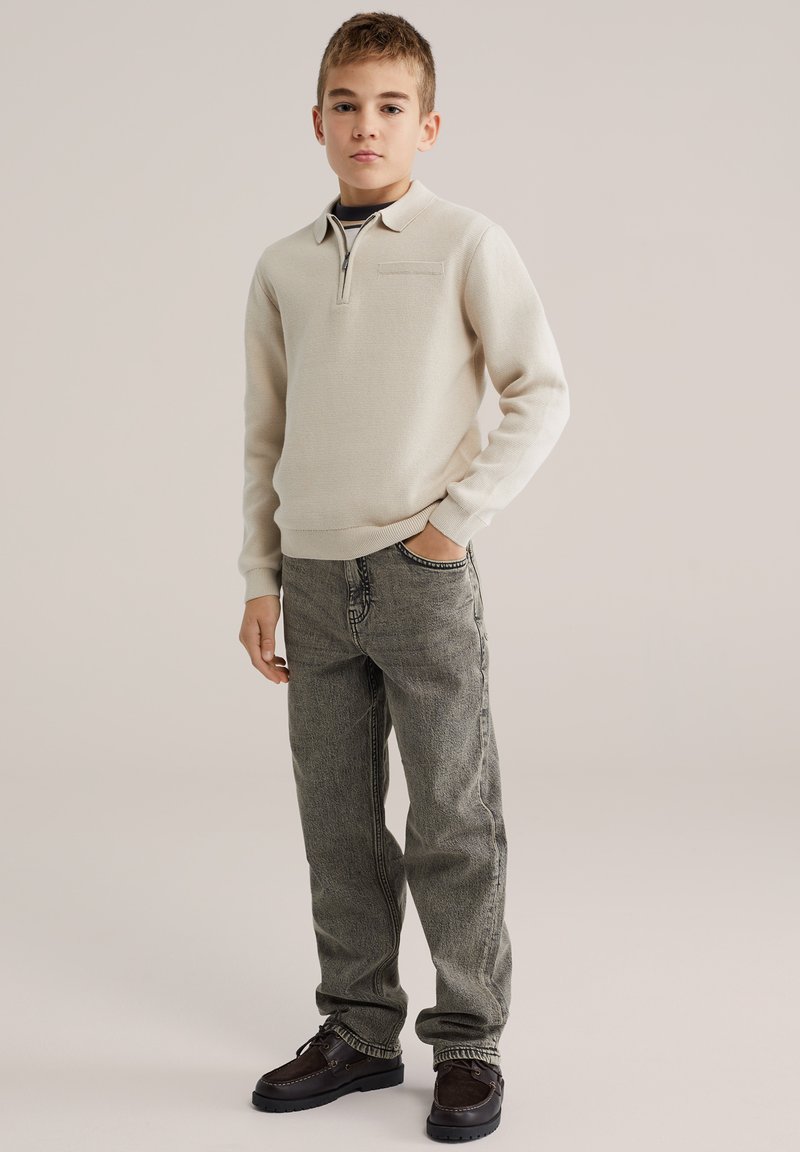 Pull beige clair avec un col zippé, tissu texturé, jeans gris et chaussures marron foncé. Le pull comporte une petite poche poitrine.