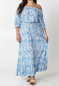 Blå axelbandslös maxiklänning med vita blommiga och bladvärden, elastisk midja och volangkant. Modell som bär beiga sandaler.