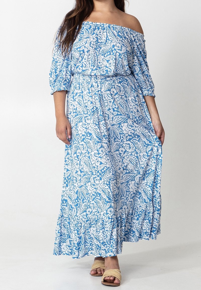 Blå axelbandslös maxiklänning med vita blommiga och bladvärden, elastisk midja och volangkant. Modell som bär beiga sandaler.