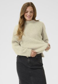 Woman with long light brown hair smiling, wearing a beige ribbed sweater and a high-waisted black denim skirt, holding the sweater hem.
