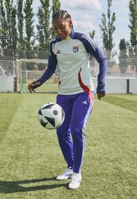 Chándal Adidas en blanco, púrpura y rojo; jugador malabareando un balón de fútbol blanco y negro sobre césped verde. Cabello trenzado y zapatos blancos.