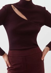 Burgundy ribbed turtleneck-paita, jossa on epäsymmetrinen leikkaus ja pitkät hihat, yhdistettynä samanvärisiin korkeavyötäröisiin housuihin.