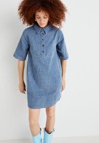 Robe bleue texturée avec un col, des manches courtes et cinq boutons à l'avant. Portée avec des bottes jusqu'aux genoux bleu clair.