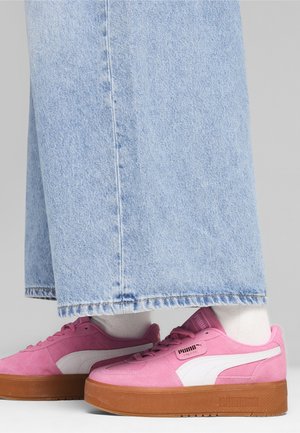 Vaaleanpunaiset Puma-lenkkarit, joissa on valkoinen raita ja kumipohjat, yhdistettynä valkoisiin sukkiin ja vaaleansinisiin leveälahkeisiin farkkuihin harmaata taustaa vasten.