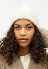 Berretto lavorato a maglia bianco con una parte superiore a punta, caratterizzato da una consistenza morbida e un motivo a coste. Il modello ha i capelli ricci e indossa un cappotto beige.