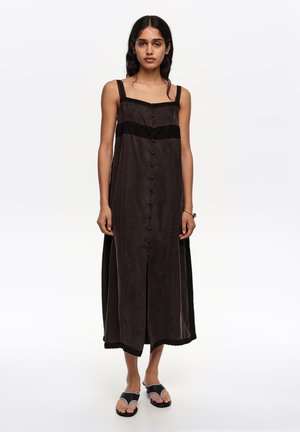 Kobieta ubrana w czarną, bezrękawnikową sukienkę midi z guzikami z przodu i czarne sandały z odkrytymi palcami, stojąca na tle białego tła.