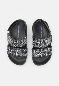 Černé sandály s dvěma nastavitelnými popruhy, na kterých je bílý text "Calvin Klein Jeans". Hladký povrch a zaoblený design špičky. Platonová podrážka.