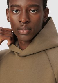 Mężczyzna o ciemnej karnacji, wearing jasnobrązowy hoodie, trzymający kaptur blisko szyi, patrząc bezpośrednio przed siebie z neutralnym wyrazem twarzy.