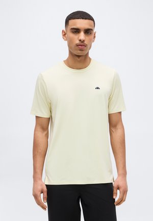 Mlad moški s kratkimi lasmi, oblečen v svetlo bež kratko majico s kratkimi rokavi in črne hlače, stoji pred enostavnim belim ozadjem.