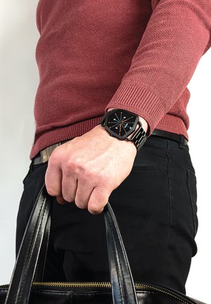 Sorte metalurisk ur med firkantet urskive, hvide og orange markeringer, båret på en hånd der holder en taske i sort læder; kastanjefarvet struktureret sweater synlig.