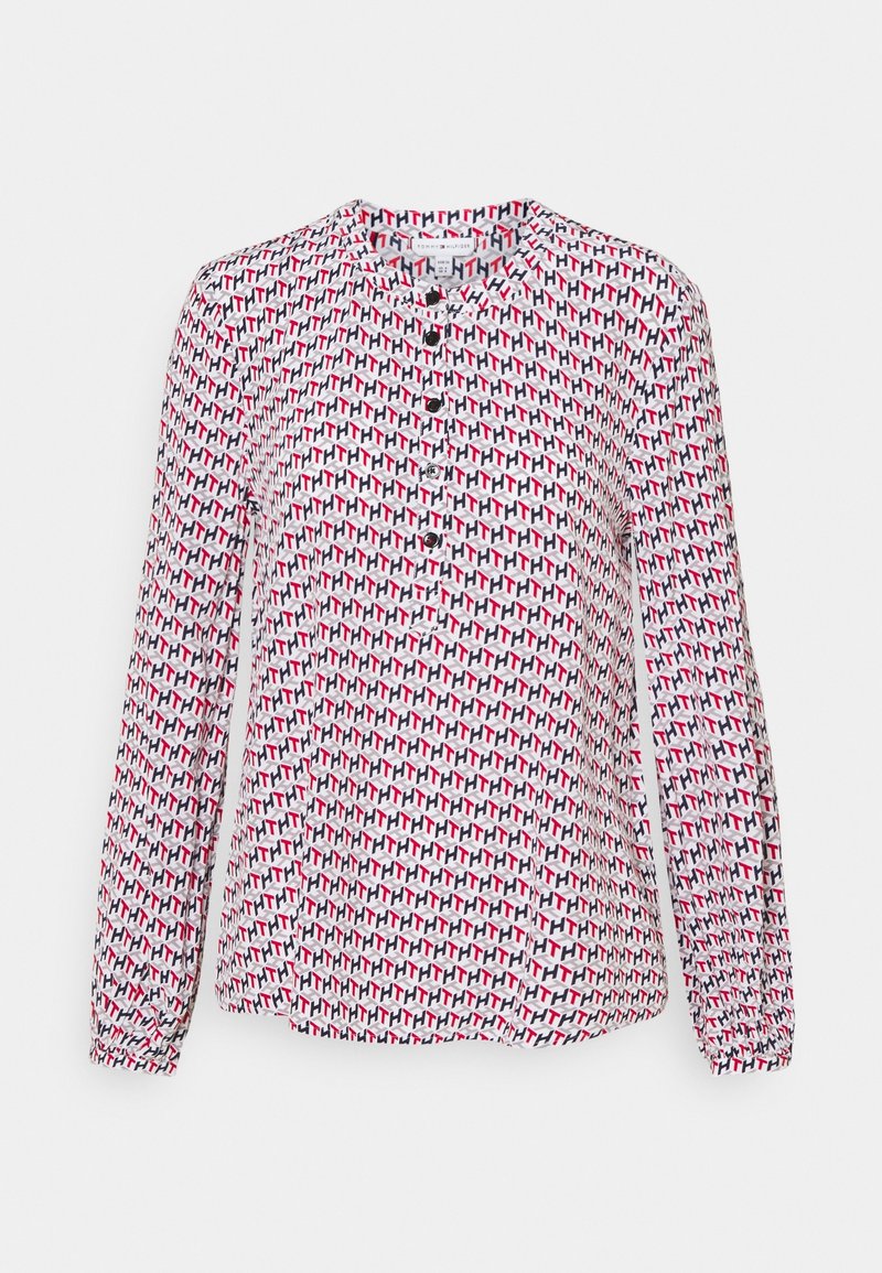 Blouse blanche à manches longues avec un motif géométrique rouge, noir et gris sur toute la surface, et des boutons noirs sur le devant.