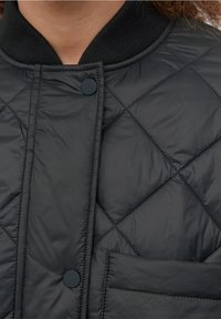 Chaqueta negra acolchada con un diseño de rombos, cuello acanalado, cierre a presión y bolsillo lateral, con una textura suave y brillante.