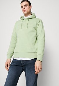 Sudadera con capucha verde claro con logo de la marca bordado. Textura suave, puños y dobladillo acanalados. Llevada sobre una camisa blanca y jeans oscuros.