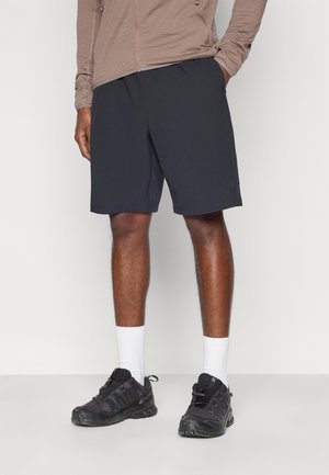 Ανδρικό σορτς σε σκουρό γκρι χρώμα από ελαφρύ ύφασμα, με ελαστική μέση. Συνδυάζεται με λευκές κάλτσες μέχρι τον αστράγαλο και μαύρα αθλητικά παπούτσια.