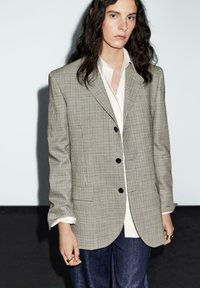 Blazer a quadri beige e marrone con bottoni neri; camicia con bottoni chiara sotto; pantaloni in denim; vestibilità ampia; maniche di lunghezza media.