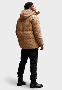 Muž v prošívané béžové bundě s kapucí, černé pletené čepici, černých cargo kalhotách a černých šněrovacích botách, stojící bokem na šedém pozadí.