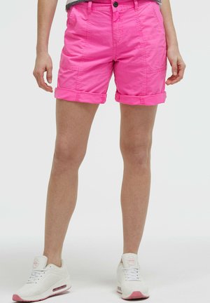 Frau trägt leuchtend pinke Shorts mit umgeschlagenem Saum und weiße Sneakers mit pinken Akzenten, steht vor einem schlichten Hintergrund.