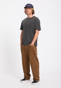 Jeune homme portant un t-shirt gris foncé, un pantalon en velours côtelé marron, des chaussures noires sans lacets et une casquette de baseball bleu marine, debout les mains dans les poches.