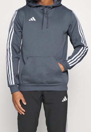 Ciemnoszara bluza z kapturem z kieszenią typu kangur, białe akcenty w postaci trzech pasków na rękawach, sznurek do regulacji kaptura oraz logo Adidas na piersi.