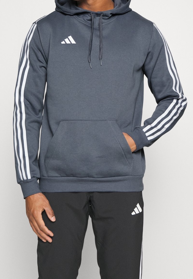 Sudadera gris oscura con bolsillo canguro, acentos en blanco con tres rayas en las mangas, capucha con cordón y logo de Adidas en el pecho.