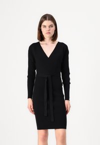 Vestido negro de malha canelada com um decote em V profundo, mangas longas e um cinto combinando na cintura. Silhueta ajustada; tecido texturizado.