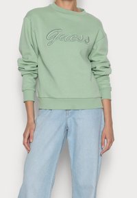 Grön sweatshirt med rund halsringning och långa ärmar, med upphöjd "Guess"-logotyp i kontrasterande färg, ihop med ljusblå vida jeans.