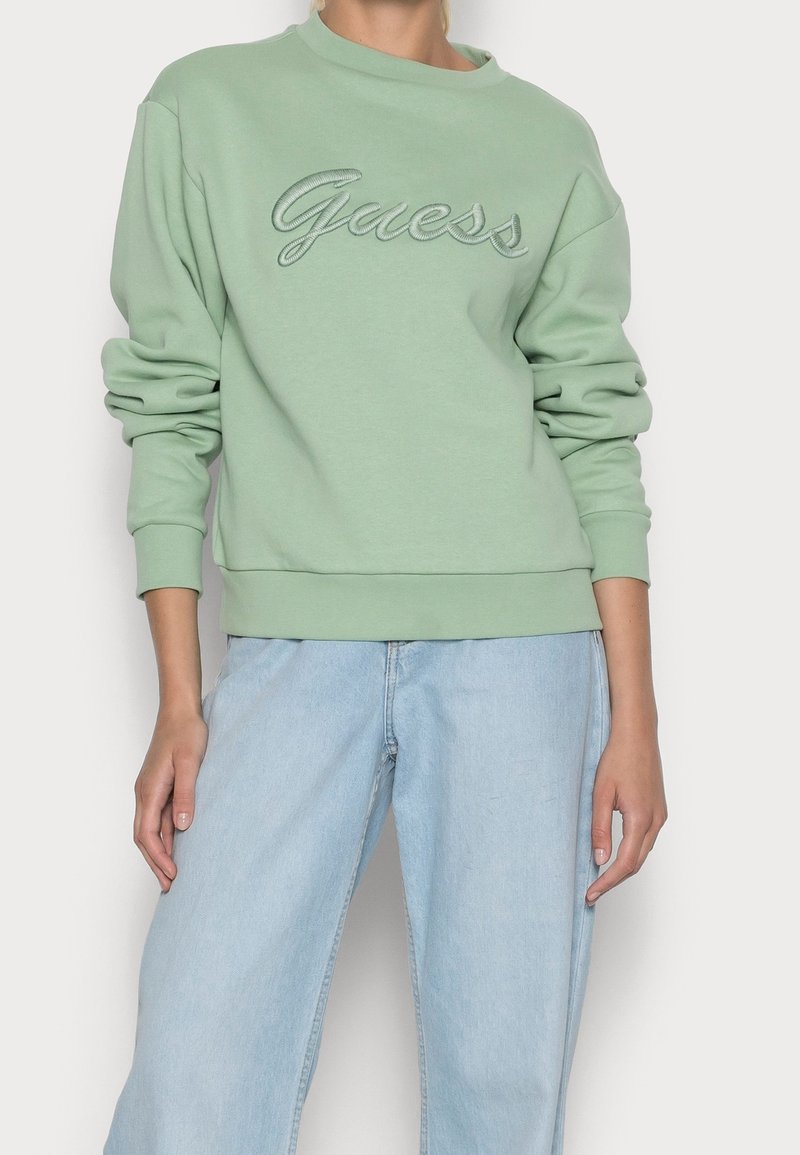 Grön sweatshirt med rund halsringning och långa ärmar, med upphöjd "Guess"-logotyp i kontrasterande färg, ihop med ljusblå vida jeans.