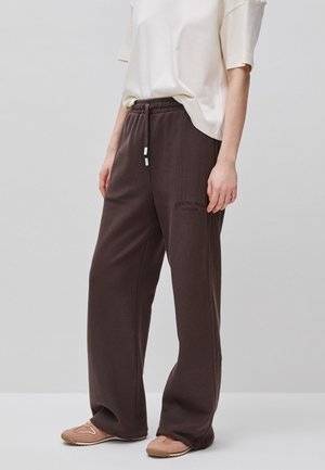 Persoon draagt losse donkerbruine joggingbroek met trekkoord en beige casual schoenen, gecombineerd met een kortemouwige off-white top.