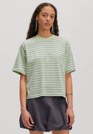 Femme portant une chemise à manches courtes rayée vert clair et une jupe ballon foncée, avec de longues boucles d'oreilles et un bracelet, debout devant un fond uni.
