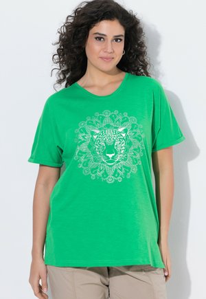 Kobieta z kręconymi włosami w zielonym T-shircie z białą głową lamparta i geometrycznym wzorem z przodu, w połączeniu z beżowymi spodniami.