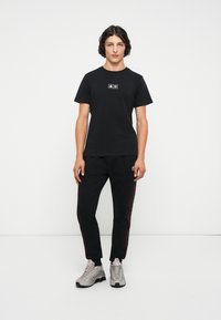 Camiseta negra de cuello redondo con parche de logo, combinada con pantalones jogger negros con franjas laterales rojas y zapatillas deportivas grises.