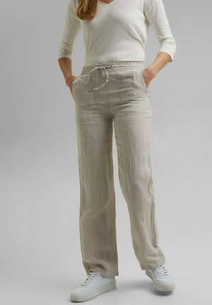 Femme portant un pantalon en lin beige à cordon, des baskets blanches et un haut crème clair à manches trois-quarts, debout les mains dans les poches.