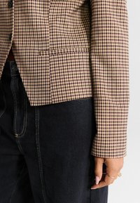 Sacou cu carouri maro și crem, cu un design ajustat, rever înclinat și detalii subtile la buzunare, purtat peste pantaloni din denim închis.