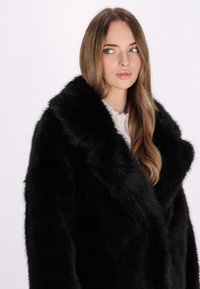 Sort faux fur frakke med en bred krave, der har en blød tekstur og en løs pasform, båret over en lysfarvet top.