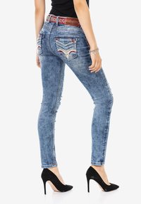 Schmale blaue Jeans mit Distressed-Details, dekorativen Gesäßtaschen und Kontrastnähten, kombiniert mit schwarzen High Heels.