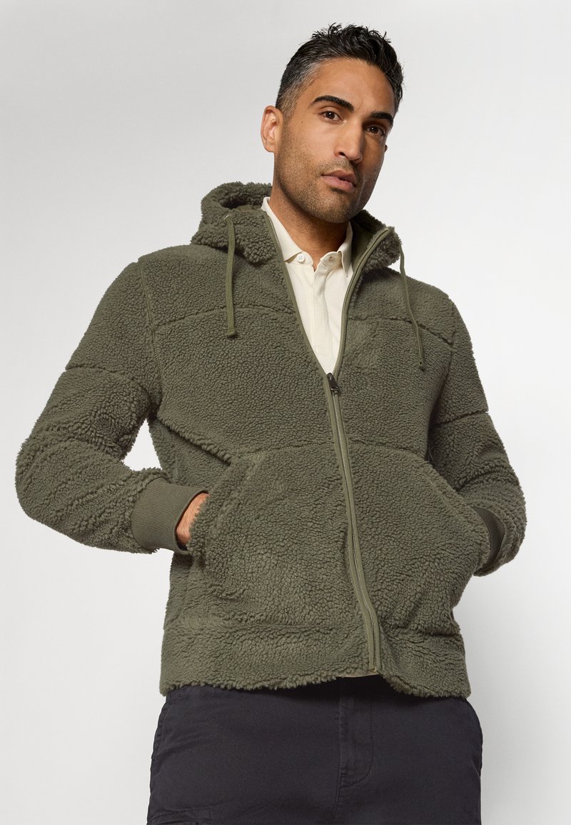 Mann trägt eine olivgrüne Fleecejacke mit Reißverschluss und Kapuze, die Hände in den vorderen Taschen, kombiniert mit dunklen Hosen und einem hellen, kragenlosen Hemd.