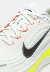 Sneaker blanc avec tissu texturé, logo swoosh noir et semelle intermédiaire jaune fluo. Présente des lacets blancs et des accents orange sur le côté.