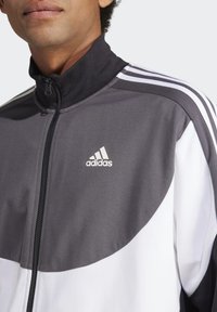 Giacca da track Adidas con colletto alto, blocchi di colore grigio e bianco, chiusura con zip e tre strisce bianche sulle spalle.