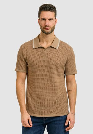 Uomo con capelli corti e barba che indossa una polo a maniche corte beige in maglia e jeans blu, in piedi davanti a uno sfondo chiaro e uniforme.