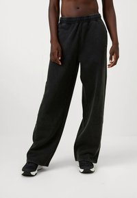 Fekete, széles szárú nadrág puha anyagból, rugalmas derékpánttal. A nadrág sima textúrájú, fekete tornacipővel viselve.