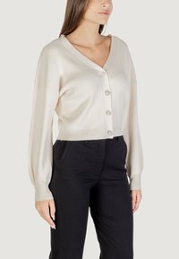 Cárdigan de canalé crema con escote en V, cinco botones con acentos de joyas y mangas largas. Combinado con pantalones negros ajustados.