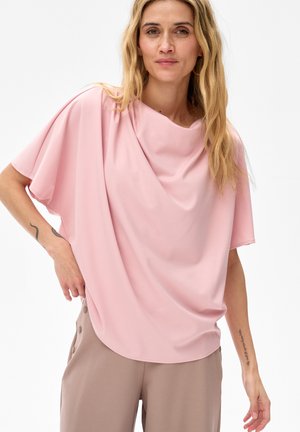 Kobieta w luźnej, różowej bluzce z krótkim rękawem i beżowych spodniach z guzikami, stojąca na tle jednolitego białego tła.